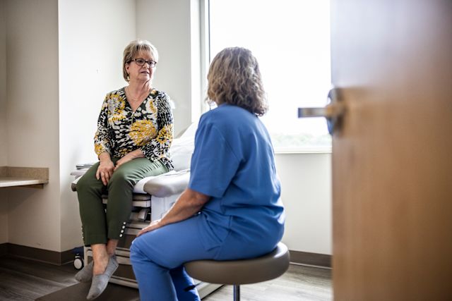 An older white woman sits on an examination table in a doctor's office while a nurse speaks with her about treatment options for a urinary tract infection.