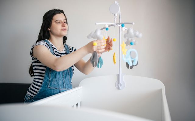 A young pregnant white woman arranges a mobile above a crib to set up a healthy non-toxic living space in anticipation of the arrival of her new baby 