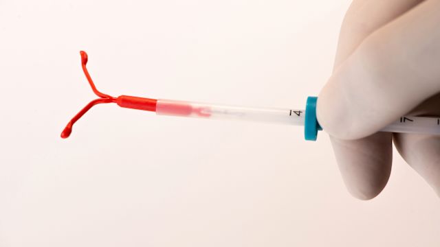A close-up of a gloved healthcare provider's hand holding a hormonal intrauterine device (IUD), a small T-shaped device made of flexible plastic.