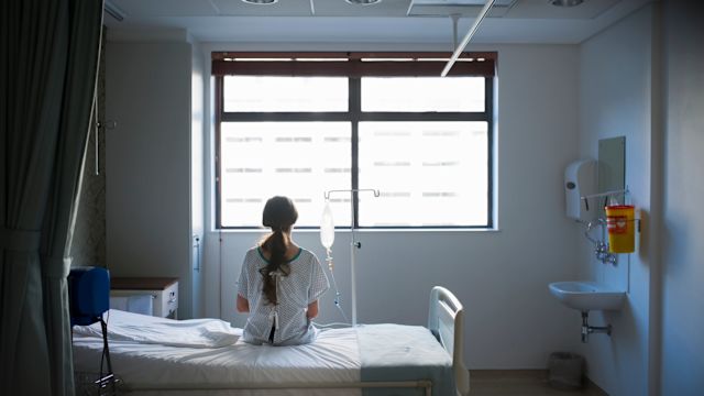A young woman with HAE sits on a hospital bed, recovering from treatment for a severe HAE attack. Preventive or prophylactic treatments can help prevent HAE attacks, but a person should also have a plan in place in case of emergencies.