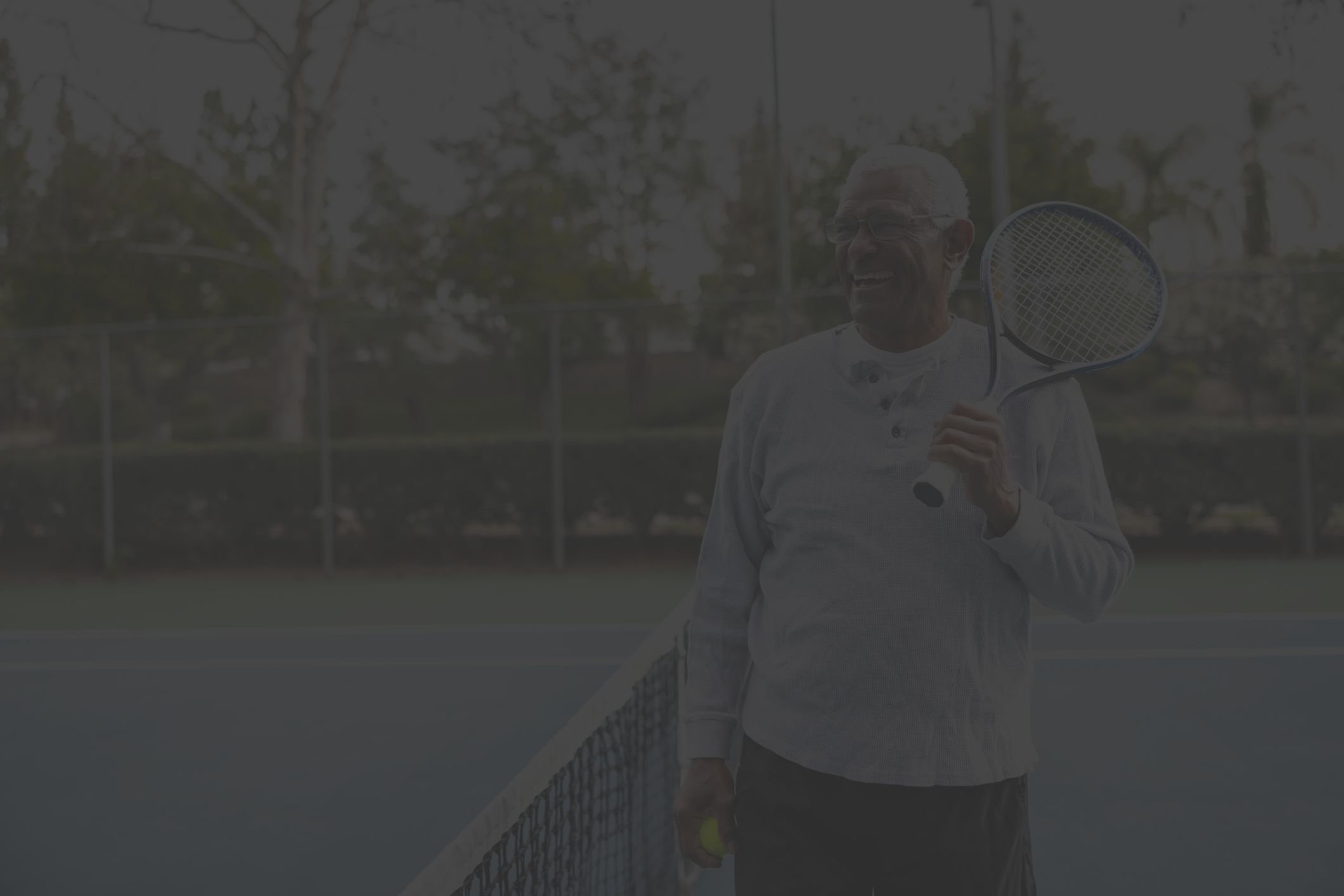 an older fit Black man stands on a tennis court holding a tennis racket and ball 