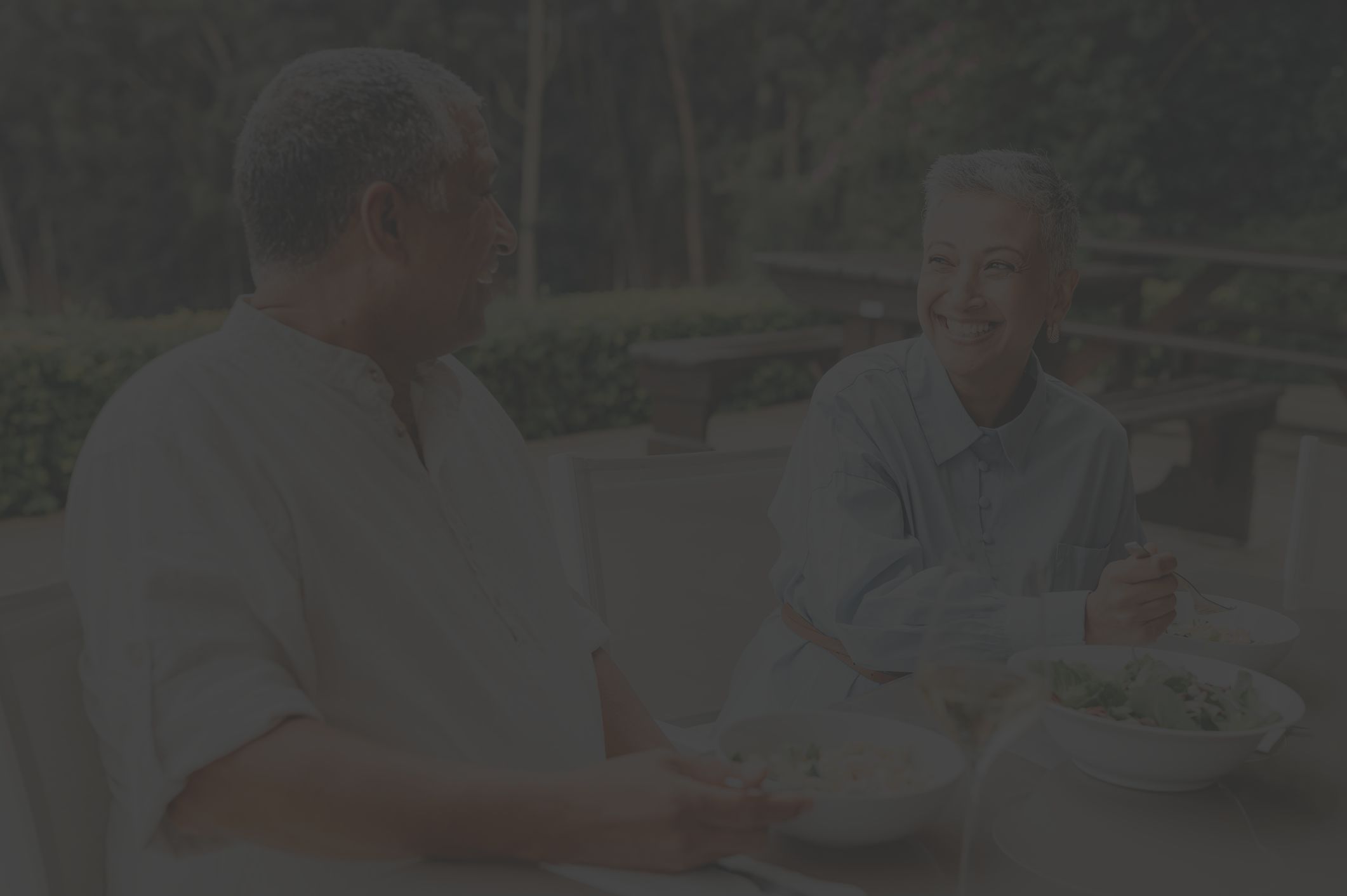 an older Latina woman with short gray hair and an older Latino man enjoy eating healthy food, including salad, in an outdoor restaurant patio