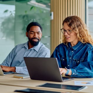 Two colleagues, a man and a woman, working together on laptops at a conference table.