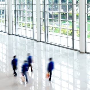 Motion-blurred figures walk across a bright, reflective marble floor in front of a wall of tall grid windows.