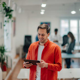 A smiling man in a bright orange shirt holds a tablet in a brightly lit office.