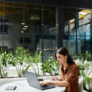 A woman with long dark hair typing on a laptop at a table surrounded by indoor plants.