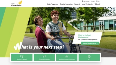 Two smiling students ride a bicycle together on a university campus path.